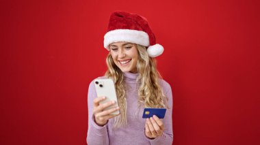 Akıllı telefon ve kredi kartıyla alışveriş yapan genç sarışın kadın izole edilmiş kırmızı arka planda noel şapkası takıyor.