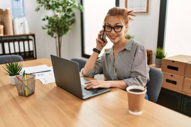 Kafkasyalı genç bir iş adamı bilgisayarını kullanarak ofisteki akıllı telefondan konuşuyor.