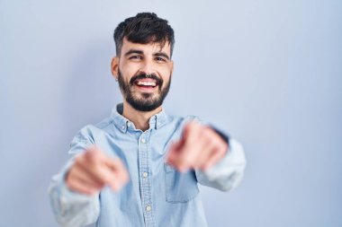 Mavi arka planda sakallı genç İspanyol adam seni ve kamerayı işaret ediyor, gülümsüyor, pozitif ve neşeli. 