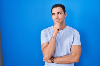 Mavi arka planda, elleri çenesinde soru soran, düşünceli bir İspanyol genç. Gülümseyen ve düşünceli bir yüz. şüphe kavramı. 