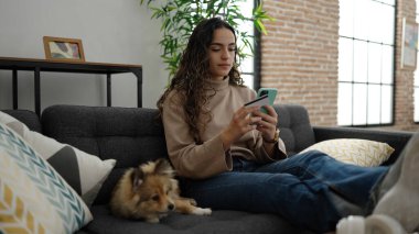 Akıllı telefon ve kredi kartıyla köpek alışverişi yapan genç İspanyol kadın evdeki kanepede oturuyor.