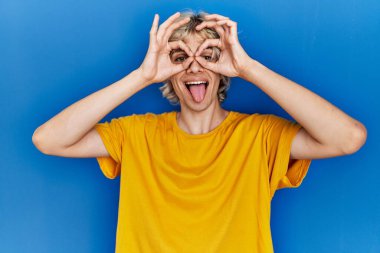 Mavi arka planda duran genç modern bir adam dürbün gibi jest yapıyor. Dürbün dilini çıkarıyor, gözler parmaklarından bakıyor. çılgın ifade. 