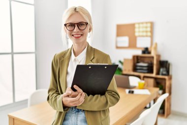 Young blonde woman business worker smiling confident holding clipboard at office