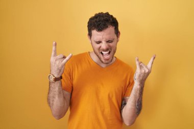 Young hispanic man standing over yellow background shouting with crazy expression doing rock symbol with hands up. music star. heavy music concept. 