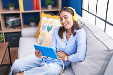 Young beautiful hispanic woman watching video on touchpad sitting on sofa at home