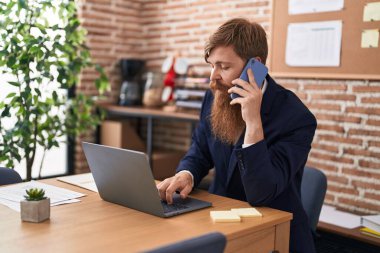 Young redhead man business worker using laptop talking on smartphone at office