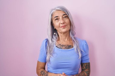 Pembe arka planda elleri çapraz parmaklı, rahat ve neşeli gülümseyen orta yaşlı bir kadın. Başarı ve iyimserlik 