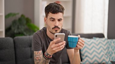 Genç İspanyol adam akıllı telefon kullanıyor. Evde sürpriz bir ifadeyle kahve içiyor.