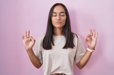 Young hispanic woman standing over pink background relaxed and smiling with eyes closed doing meditation gesture with fingers. yoga concept. 