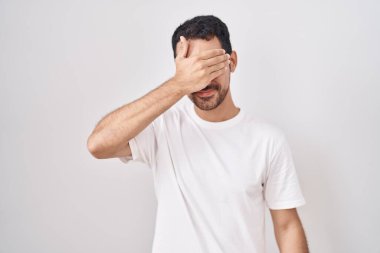 Beyaz arka planda duran yakışıklı İspanyol adam. Gülümsüyor ve gülümsüyor. Gözlerini şaşkınlıkla kapatıyor. kör kavram. 