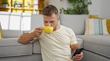 Genç adam akıllı telefon kullanıyor. Evde kahve içiyor.