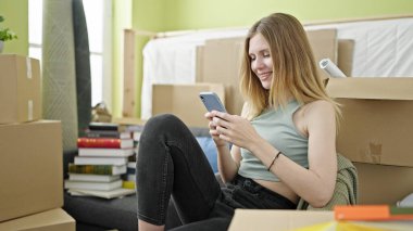 Young blonde woman using smartphone sitting on sofa at new home
