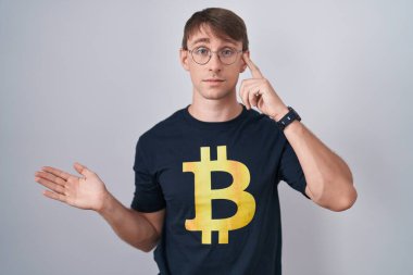 Bitcoin tişörtü giyen beyaz, sarışın bir adam açık avuç içinin fotokopi alanını göstermesi ve alnını işaret etmesi kafasını karıştırmış ve rahatsız etmiş. Bunu bir düşün.. 