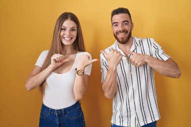 Sarı arka planda duran genç çift, elleri ve başparmakları havada, kendinden emin bir şekilde sırıtıyor. 