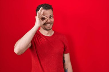 Kırmızı arka planda dikilen İspanyol genç adam el hareketleriyle gülümsüyor ve mutlu yüzüyle parmaklarının arasından bakıyor.. 
