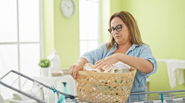 Orta yaşlı İspanyol kadın çamaşır odasında çamaşır sepetinde kıyafet arıyor.
