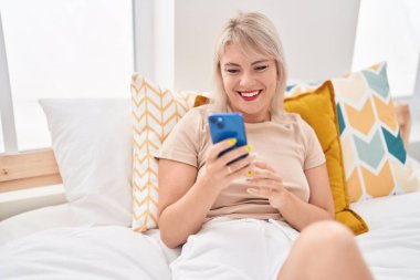 Yatak odasında akıllı telefon kullanan genç sarışın kadın.