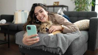 Köpekli İspanyol bir kadın akıllı telefonuyla evdeki kanepede selfie çekiyor.