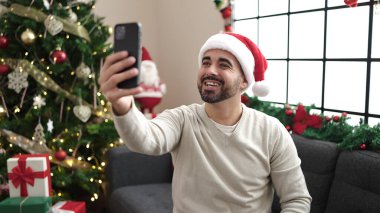 Genç İspanyol adam akıllı telefonuyla selfie çekiyor. Evdeki Noel ağacının yanında, koltukta oturuyor.