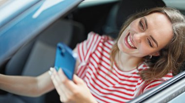 Genç sarışın kadın akıllı telefon kullanıyor. Sokakta arabada oturuyor.
