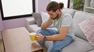 Genç İspanyol adam evdeki kanepede kahve içiyor.