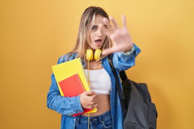 Öğrenci çantası takan ve kitap tutan genç sarışın kadın avuç içi hareketlerini durduruyor, kızgın ve hayal kırıklığına uğramış bir ifade takınıyor. 