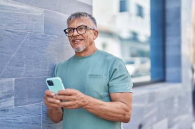Orta yaşlı, kır saçlı, kendine güvenen, caddede akıllı telefon kullanan bir adam.