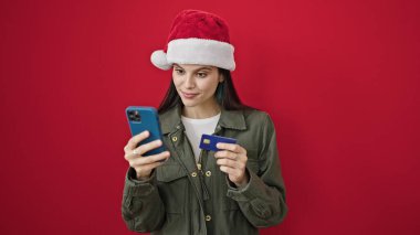 Akıllı telefon ve kredi kartıyla alışveriş yapan genç ve güzel İspanyol kadın izole edilmiş kırmızı arka planda noel şapkası takıyor.