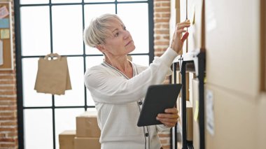 Orta yaşlı sarışın kadın ecommerce iş işçisi ofiste dokunmatik ped kullanarak paketleri kontrol ediyor.