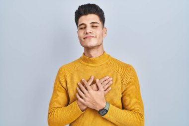 Mavi arka planda duran genç İspanyol adam elleri göğsünde, gözleri kapalı ve yüzünde minnet dolu bir jest ile gülümsüyor. sağlık kavramı. 