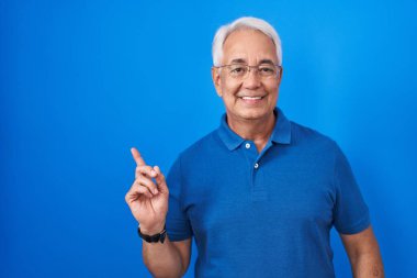 Mavi arka planda gri saçlı, yüzünde kocaman bir gülümseme olan orta yaşlı bir adam, eli ve parmağı kameraya bakıyor.. 