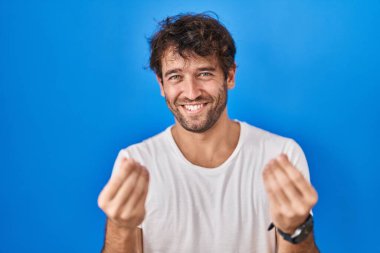 Mavi arka planda duran İspanyol genç adam elleriyle para hareketi yapıyor, maaş istiyor, milyonerlik işi yapıyor. 