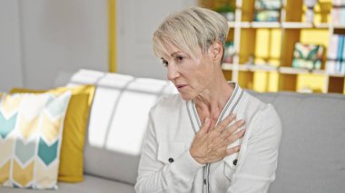 Kanepede oturan orta yaşlı sarışın kadın evinde kalp krizi geçiriyor.