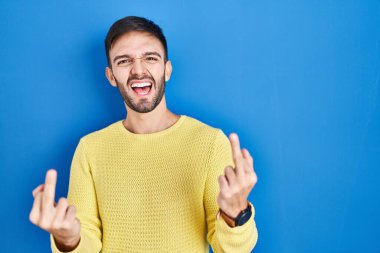 Hispanic man standing over blue background showing middle finger doing fuck you bad expression, provocation and rude attitude. screaming excited 