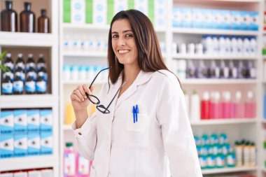 Young beautiful hispanic woman pharmacist smiling confident holding glasses at pharmacy