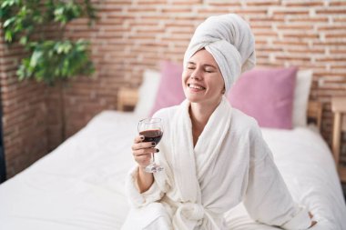 Genç sarışın kadın bornoz giyiyor. Yatak odasında şarap içiyor.