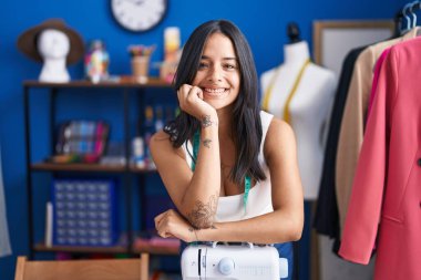 Young hispanic woman tailor smiling confident leaning on sewing maching at sewing studio