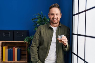 Genç beyaz adam evde dikilip bir bardak viski içiyor.