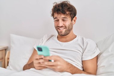 Young man using smartphone sitting on bed at bedroom