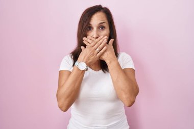 Orta yaşlı esmer kadın pembe arka planda dikiliyor. Ağzını yanlışlıkla elleriyle kapatıyor. gizli kavram. 