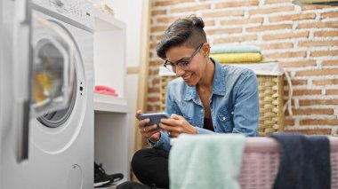 Young beautiful hispanic woman using smartphone waiting for washing machine at laundry room