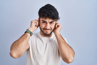 Hispanic man with beard standing over white background covering ears with fingers with annoyed expression for the noise of loud music. deaf concept. 
