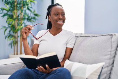 African american woman reading book sitting on sofa at home