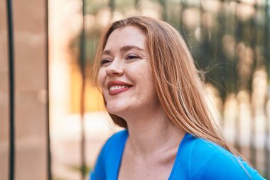 Genç kızıl saçlı kadın gülümsüyor. Kendinden emin bir şekilde caddenin kenarına bakıyor.