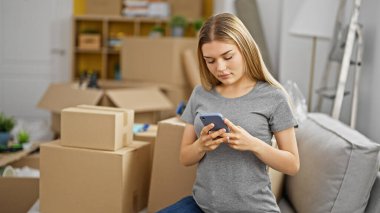 Young blonde woman using smartphone sitting on sofa at new home