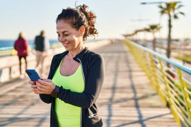 Middle age hispanic woman working out with smartphone at promenade