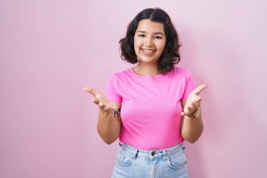 Young hispanic woman standing over pink background smiling cheerful offering hands giving assistance and acceptance. 