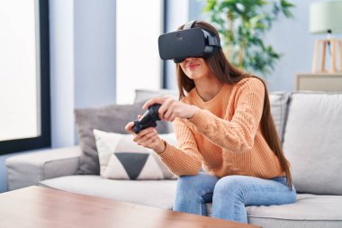 Evde sanal gerçeklik gözlüğü ve joystick kullanan güzel İspanyol bir kadın oyun oynuyor.