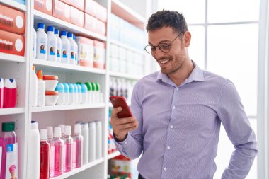 Genç İspanyol adam eczanede akıllı telefon kullanarak gülümsüyor.