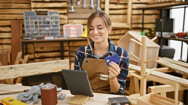 Attractive young blonde woman carpenter smiling while paying with credit card on touchpad amidst carpentry workshop